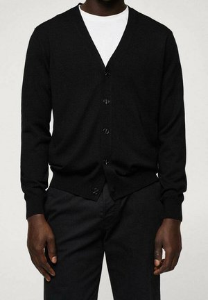 Uomo che indossa un cardigan nero abbottonato sopra una camicia bianca e pantaloni neri, in piedi contro uno sfondo chiaro uniforme.
