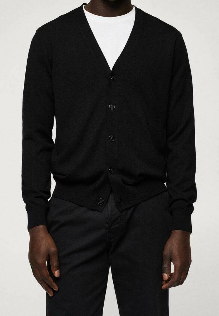 Homme portant un cardigan noir boutonné sur une chemise blanche et un pantalon noir, debout devant un fond clair uni.