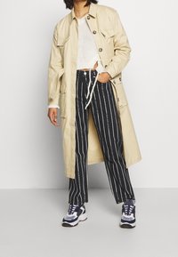 Beige lång kappa med stora knappar över en vit tröja. Svartvita randiga byxor med logotyper. Skor: marinblåa och vita sneakers.