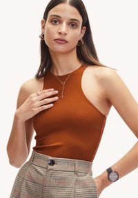 Roestbruine tanktop met een hoge halslijn, gestructureerde stof en een open schouderontwerp, gecombineerd met een geruit broek en zilveren sieraden accenten.