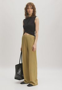 Zwarte mouwloze top met een gedraaid ontwerp, gecombineerd met losse, gestructureerde olijfgroene broek. Het model houdt een zwarte tote bag vast en draagt platte zwarte schoenen.
