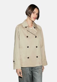 Trench coat beige doppiopetto con bottoni marroni, tasche laterali e una vestibilità rilassata. Il tessuto ha una trama liscia.