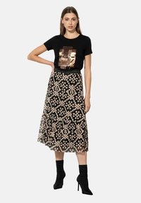 Zwarte T-shirt met metallic grafiek, gecombineerd met een doorzichtige bloemenrok met een gelaagde zoom. Model draagt zwarte hakken.