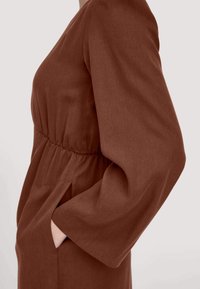 Braunes Kleid mit einem taillierten Bund, langen, voluminösen Ärmeln und seitlichen Taschen. Hergestellt aus strukturiertem Stoff, zeichnet sich durch eine schlichte, enganliegende Silhouette aus.