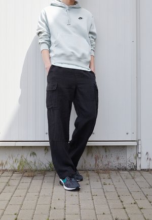 Personne portant un sweat à capuche Nike gris clair, un pantalon cargo noir et des baskets noires et turquoise, debout les mains dans les poches.