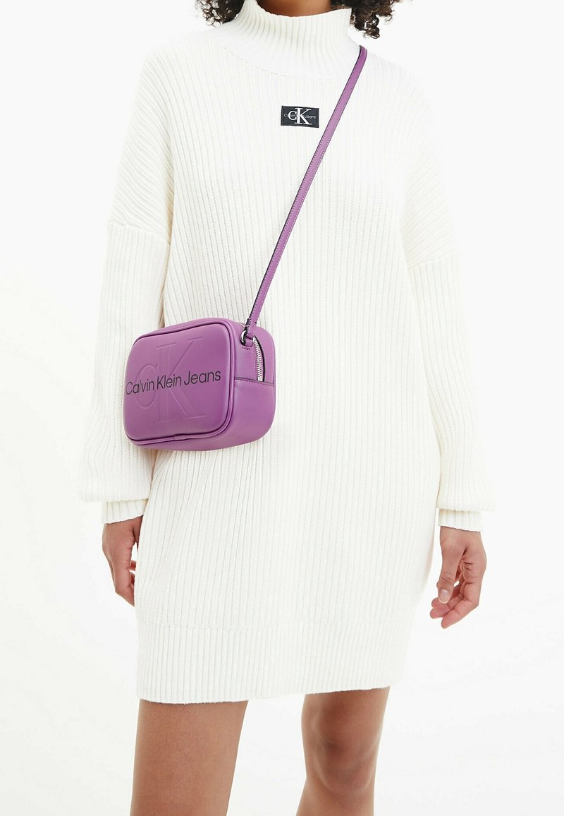 Robe pull blanche à côtes avec un col haut et un patch logo noir. Sac bandoulière violet avec logo en relief et fermeture éclair.