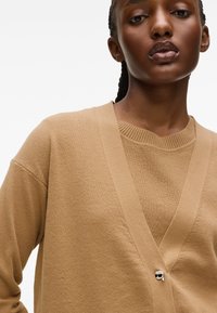 Beige kardigán V-nyakkal és oversize szabással, jellegzetes ezüst koponya részletekkel. Puha, texturált kötött anyag.