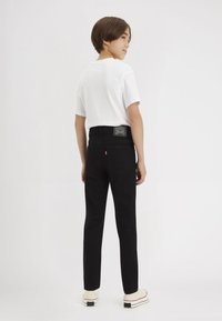 Fekete farmernadrág slim fit fazonban, hátsó zsebekkel, bőr címkével, fehér pólóval és világosbézs sportcipővel párosítva.