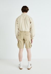 Personne aux cheveux courts et bouclés portant une chemise beige à manches longues, un short cargo beige, des chaussettes blanches et des chaussures blanches, debout de dos.