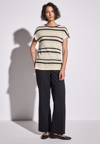 Short-sleeve top in cream with black patterned stripes, paired with loose black trousers and black flats. Smooth fabric with a relaxed fit.