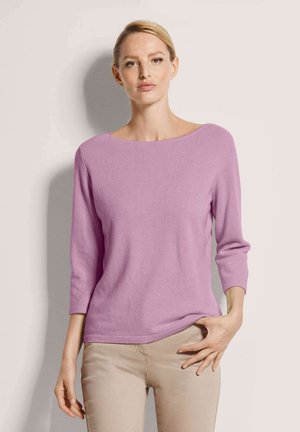 Frau mit blonden Haaren, die einen lila Pullover mit Dreiviertelärmeln und beigen Hosen trägt, steht vor einem hellen Hintergrund.