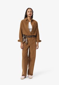 Camicia di corduroy marrone e pantaloni a gamba larga coordinati, abbinati a una cintura con motivo e sneakers di colore chiaro. Texture morbida, vestibilità rilassata.