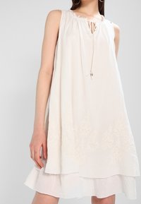 Ärmelloses, lockeres weißes Kleid mit dezenten Blumenstickereien am Saum und einer Bindung am Ausschnitt, getragen von einer stehenden Frau.