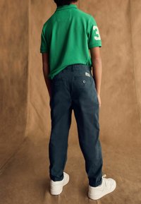 Groene polo met witte nummer 3, gecombineerd met marineblauwe broek. De achtergrond is een glad, neutraal bruin. Witte sneakers zijn zichtbaar.