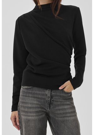 Femme portant un haut noir à manches longues avec des plis diagonaux et un jean gris, debout avec une main dans la poche devant un fond uni.