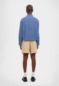 Ljus denimskjorta med långa ärmar, en spetsig krage och rak nederkant, i kombination med beiga shorts. Inkluderar knäppbara manschetter och subtil textur.