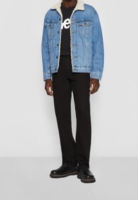 Chaqueta de mezclilla azul claro con cuello de peluche sintético, camiseta negra con texto blanco, pantalones negros de pierna recta y botas negras de suela gruesa.