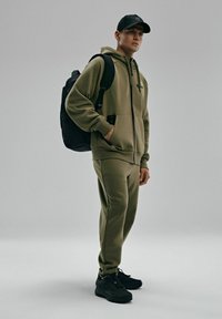 Zestaw oliwkowej zielonej bluzy i dresów wykonany z miękkiego materiału, z kapturem na zamek błyskawiczny, czarnymi akcentami oraz eleganckimi czarnymi sneakersami. W zestawie czarna plecak.