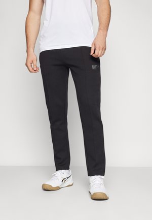 Pantaloni della tuta neri a tapering realizzati in un tessuto morbido, con una patch testurizzata sul fronte. Abbinati a una maglietta bianca e scarpe da ginnastica bianche.