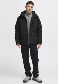 Sorte vandtætte jakke med hætte, knaplukning fortil og to forbagslommer. Båret over en grå tekstureret sweater og sorte bukser.