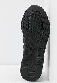 Schwarze Gummisohle eines New Balance Schuhs mit gerilltem Profil, vertikal gegen eine weiße, eckige Wand gelehnt.