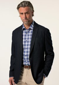 Marinblazer med en strukturerad design, kombinerad med en blå och vit rutig skjorta. Ljusbeige byxor och brunt bälte synliga.