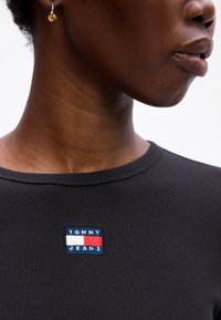 Maglietta nera a righe con un logo ricamato rettangolare "TOMMY JEANS" blu, bianco e rosso nella zona sinistra del petto.