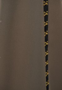 Brunt stof med en glat tekstur prydes af en sort sidelæbe, der er udsmykket med gyldne dekorative hardware detaljer.