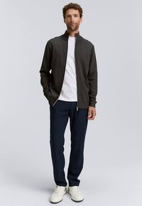 Pilko spalvos džemperis su užtrauktuku per vidurį dėvimas ant baltos palaidinės, derinamas su tamsiai mėlynais kelnėmis ir baltais batais. Audinys atrodo glotnus, o drabužis – laisvas ir kasdieniškas.