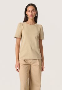 Beige bomulds t-shirt med korte puffærmer og rund halsudskæring, parret med matchende beige bukser, præsenteret mod en neutral baggrund.