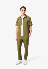 Camisa verde de botones de manga corta con una camiseta blanca debajo, combinada con pantalones verdes que presentan un sutil patrón de logotipos repetidos. Zapatos blancos.