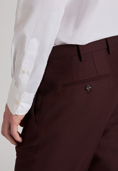 Camisa blanca de manga larga con puños con botones, combinada con pantalones a medida de color marrón oscuro que cuentan con un bolsillo con cierre de botón.