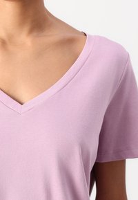 Detailní záběr na osobu v světle fialovém tričku s výstřihem do V a krátkým rukávem na jednoduchém bílém pozadí.