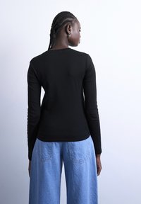 Γυναίκα με πλεγμένα μαλλιά που φοράει μαύρο μπλουζάκι με μακριά μανίκια και ανοιχτό μπλε τζιν με φαρδιά μπατζάκια, γυρισμένη πλάτη σε λευκό απλό φόντο.