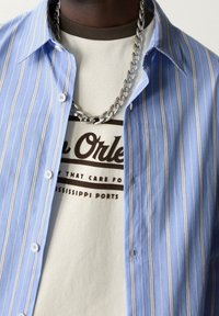 Chemise à rayures bleu clair avec boutons, portée sur un t-shirt graphique beige avec texte noir. Collier chaîne argenté visible.