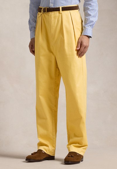 Pantalon plissé jaune en coton, avec une coupe décontractée, associé à une ceinture marron et des mocassins marrons. Chemise rayée bleu et blanc.