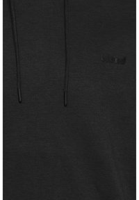 Sudadera negra con un tejido texturizado, que cuenta con dos cordones y un pequeño logo negro en la parte delantera. Diseño simple sin patrones.