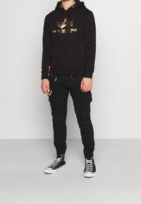 Svart hoodie med guldlogga, kombinerad med svarta cargobyxor med dragkedjor och fickor. Bärs med svarta högskor.