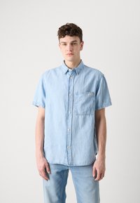 Camisa de mezclilla de manga corta azul claro con cierre de botones, cuello y bolsillo en el pecho. Textura suave con un ajuste relajado.