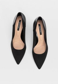 Zapatos negros de punta con un diseño elegante, que presentan una combinación de cuero suave y tejido blando, mostrando un detalle recortado en el costado.