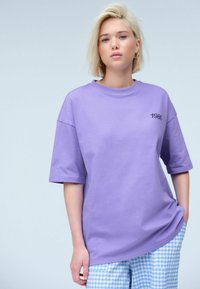 Lila Baumwoll-T-Shirt mit kurzen Ärmeln und Rundhalsausschnitt, auf der Vorderseite mit dem Text "1980" in Schwarz bedruckt. Kombiniert mit blauen Gingham-Hosen.