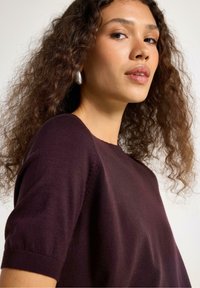 Donkerpaarse gebreide top met korte mouwen, met een ronde halslijn en een soepele textuur. Het model heeft krullend haar en draagt zilveren oorbellen.