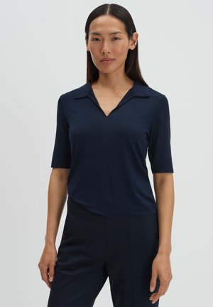 Donna con capelli lisci e neri indossa una camicia con colletto a maniche corte blu scuro e pantaloni abbinati su uno sfondo chiaro e uniforme.