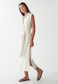 Donna che indossa un vestito midi beige senza maniche, con bottoni, cintura e tasche, che cammina in sandali piatti neri con cinturini su uno sfondo bianco.