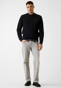 Zwarte trui met een ronde hals en subtiel logo, lichtgrijze spijkerbroek en zwarte schoenen, staand tegen een effen achtergrond.