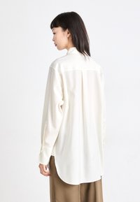 Lange mouwen, crèmekleurige blouse met een zachte textuur, achterpleatdetail en afgeronde zoom. Voorzien van knoopmanchetten en een klassieke kraag.