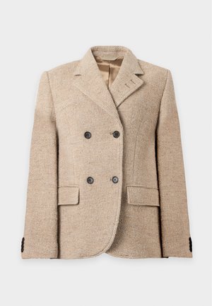 Model trägt einen beigen zweireihigen Wollblazer, ein hellblau gestreiftes Hemd, hellblaue Jeans mit weitem Bein und schwarze spitze Schuhe vor weißem Hintergrund.