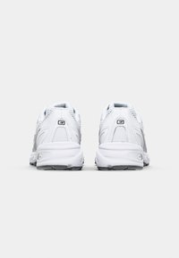 Vita idrottsskor i vitt med en strukturerad ovandel, vadderad krage och gummisula med svarta accenter. Bakifrån visas ytterligare brandingdetaljer.