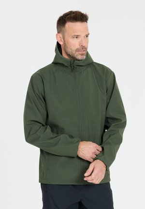 Man die mouw van een groene capuchonjas verstelt, licht naar rechts gericht, tegen een effen witte achtergrond.