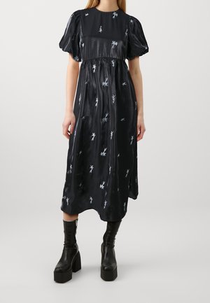 Frau trägt ein schwarzes Midikleid mit Puffärmeln und weißen Blumendesigns, kombiniert mit schwarzen Plateau-Stiefeletten mit Reißverschlüssen.